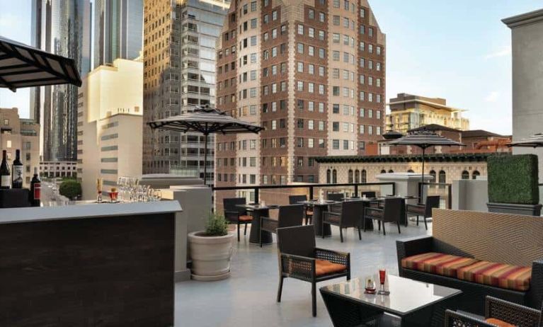 Elegant bar and a spacious rooftop filled with couches and small tables, skyline backdrop of downtown Los Angeles.