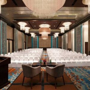 Long presentation and banquet hall with ornate chandeliers, a podium and sitting area for two panelists at the front, at the Hilton Jaipur, India.