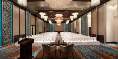 Long presentation and banquet hall with ornate chandeliers, a podium and sitting area for two panelists at the front, at the Hilton Jaipur, India.