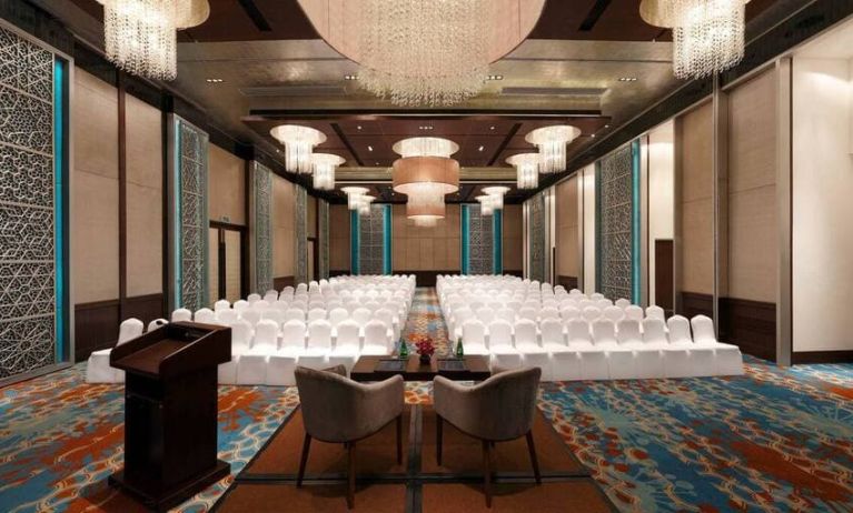 Long presentation and banquet hall with ornate chandeliers, a podium and sitting area for two panelists at the front, at the Hilton Jaipur, India.