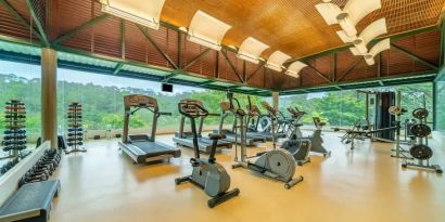 Exercise room with treadmills, ellipticals, space for free weights, with floor to ceiling windows surrounded by view of trees.