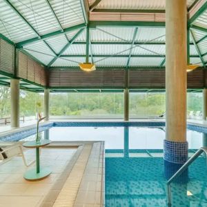 Large pillar in middle of indoor pool with floor to ceiling windows and space to lounge.