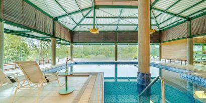 Large pillar in middle of indoor pool with floor to ceiling windows and space to lounge.