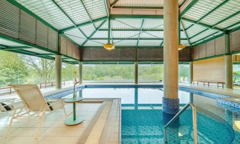 Large pillar in middle of indoor pool with floor to ceiling windows and space to lounge.