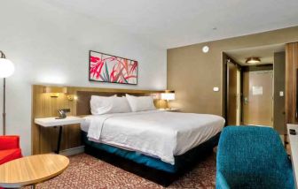 Queen size bed with art hanging over, television and blue ottoman, bright red chair with end table to sit.