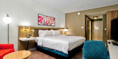 Queen size bed with art hanging over, television and blue ottoman, bright red chair with end table to sit.