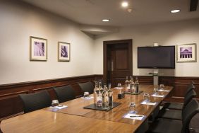 A large glossy wooden table with glass water carafes and seating for 8 participants with large television mounted on wall.