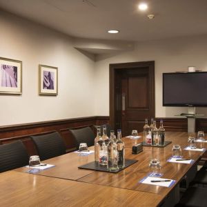 A large glossy wooden table with glass water carafes and seating for 8 participants with large television mounted on wall.