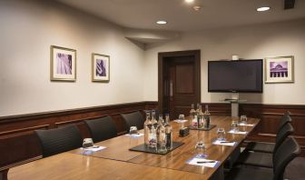 A large glossy wooden table with glass water carafes and seating for 8 participants with large television mounted on wall.