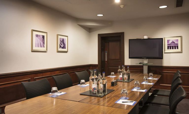 A large glossy wooden table with glass water carafes and seating for 8 participants with large television mounted on wall.