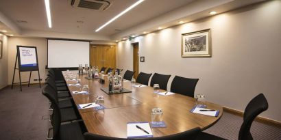 A wider shot of the meeting room with over half a dozen chairs, multiple tables, and a screen.