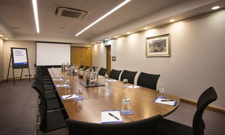 A wider shot of the meeting room with over half a dozen chairs, multiple tables, and a screen.