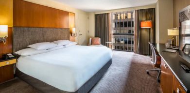 Guest room in Hyatt Regency Chicago, with large double bed, window, and workspace desk, chair, and lamp.