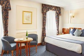 An executive double standard room at the Thistle London Heathrow Terminal 5, with large bed, windows, and coffee table and chairs.