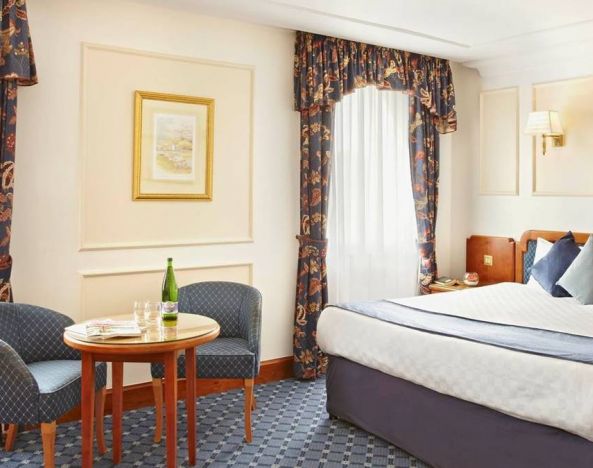 An executive double standard room at the Thistle London Heathrow Terminal 5, with large bed, windows, and coffee table and chairs.