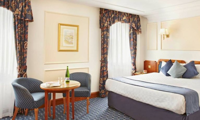 An executive double standard room at the Thistle London Heathrow Terminal 5, with large bed, windows, and coffee table and chairs.