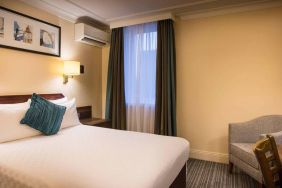 Double bed in a standard room at the Thistle London Heathrow Terminal 5, with nearby window and chair.