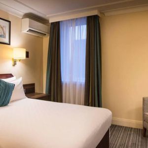 Double bed in a standard room at the Thistle London Heathrow Terminal 5, with nearby window and chair.