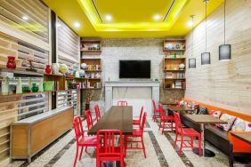 A long rectangular wooden table in the middle with red chairs. Several other smaller tables for 4 to work, eat, socialize at. Yellow lit ceiling and white fireplace mantle frames the room.