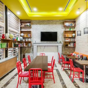 A long rectangular wooden table in the middle with red chairs. Several other smaller tables for 4 to work, eat, socialize at. Yellow lit ceiling and white fireplace mantle frames the room.