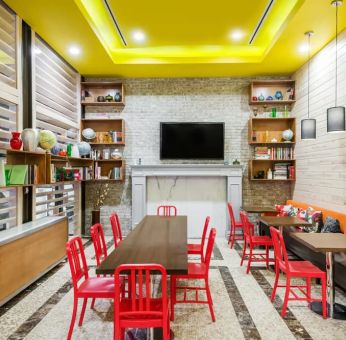 A long rectangular wooden table in the middle with red chairs. Several other smaller tables for 4 to work, eat, socialize at. Yellow lit ceiling and white fireplace mantle frames the room.