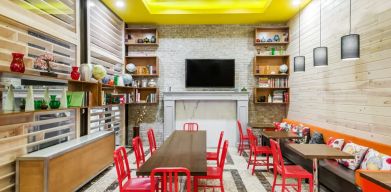 A long rectangular wooden table in the middle with red chairs. Several other smaller tables for 4 to work, eat, socialize at. Yellow lit ceiling and white fireplace mantle frames the room.