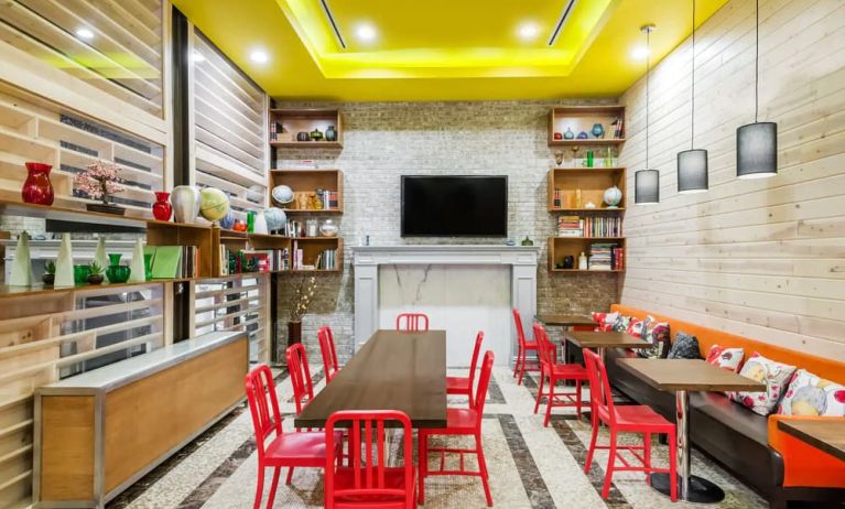 A long rectangular wooden table in the middle with red chairs. Several other smaller tables for 4 to work, eat, socialize at. Yellow lit ceiling and white fireplace mantle frames the room.