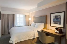 Cozy hotel room with beige curtains, wall-mounted television, white linen over full-size bed, with adjacent desk complete with coffeemaker and comfortable leather chair.