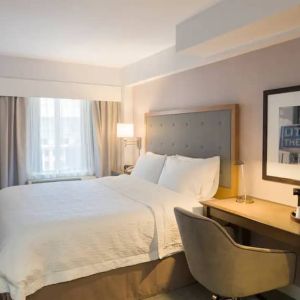 Cozy hotel room with beige curtains, wall-mounted television, white linen over full-size bed, with adjacent desk complete with coffeemaker and comfortable leather chair.