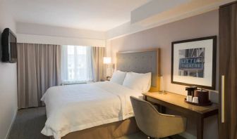 Cozy hotel room with beige curtains, wall-mounted television, white linen over full-size bed, with adjacent desk complete with coffeemaker and comfortable leather chair.