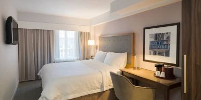 Cozy hotel room with beige curtains, wall-mounted television, white linen over full-size bed, with adjacent desk complete with coffeemaker and comfortable leather chair.