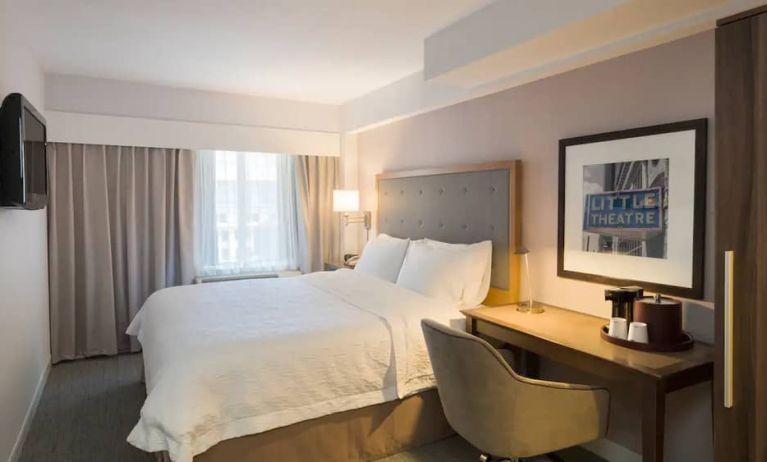 Cozy hotel room with beige curtains, wall-mounted television, white linen over full-size bed, with adjacent desk complete with coffeemaker and comfortable leather chair.