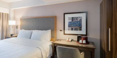 Full-size bed with olvie bed header, adjacent hotel room workspace with seude chair, coffeemaker, phone and desk lamp.