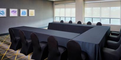 Hotel meeting room with three tables arranged in a U-shape and seating for over a dozen attendees.