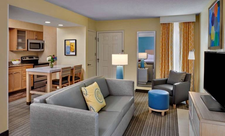 Sonesta ES Suites Cincinnati - Blue Ash guest room lounge, with armchair, sofa, and television, plus nearby kitchenette.