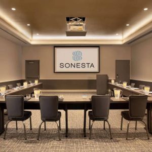 Hotel meeting room with long tables arranged in a U-shape, facing a large projector screen.