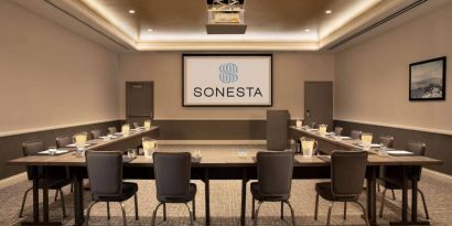 Hotel meeting room with long tables arranged in a U-shape, facing a large projector screen.