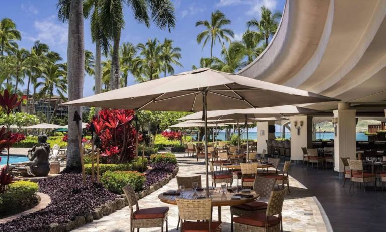 The hotel’s terrace includes dining under shaded cover, with the pool and towering trees nearby.