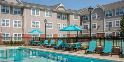 The outdoor pool at Sonesta ES Suites Charlottesville University has a mix of sun loungers and shaded tables and chairs by the side.