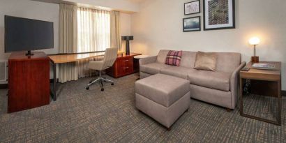 Sonesta ES Suites Baltimore BWI Airport guest room workspace, featuring chair, desk, and TV, plus a sofa nearby.