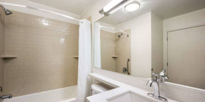 Guest bathroom in Sonesta Simply Suites Des Moines, including a long mirror, lavatory, sink, and bath with a shower.