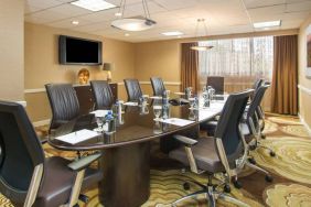 Hotel meeting room, furnished with long table, 10 swivel chairs, chocolate hues, and a wall-mounted television.