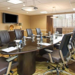 Hotel meeting room, furnished with long table, 10 swivel chairs, chocolate hues, and a wall-mounted television.