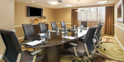 Hotel meeting room, furnished with long table, 10 swivel chairs, chocolate hues, and a wall-mounted television.