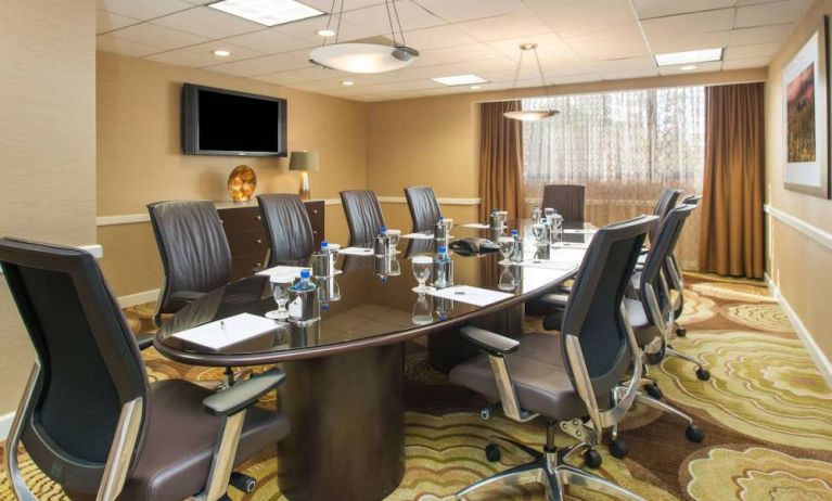 Hotel meeting room, furnished with long table, 10 swivel chairs, chocolate hues, and a wall-mounted television.