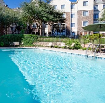 Sonesta ES Suites Austin The Domain Area’s outdoor pool features tables and chairs by the side, some of which are shaded, and nearby trees.