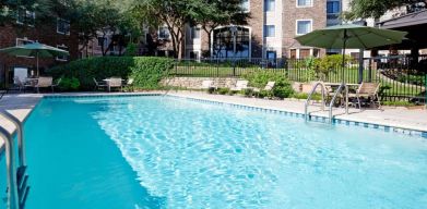 Sonesta ES Suites Austin The Domain Area’s outdoor pool features tables and chairs by the side, some of which are shaded, and nearby trees.