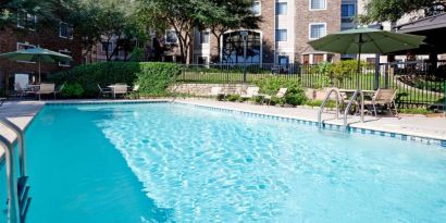 Sonesta ES Suites Austin The Domain Area’s outdoor pool features tables and chairs by the side, some of which are shaded, and nearby trees.