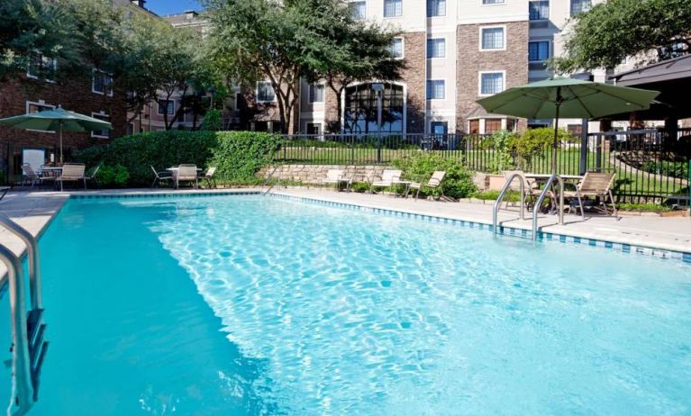 Sonesta ES Suites Austin The Domain Area’s outdoor pool features tables and chairs by the side, some of which are shaded, and nearby trees.