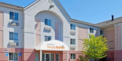 Sonesta Simply Suites Detroit Ann Arbor’s exterior features clear signage, and a pair of benches beside foliage including a tree.